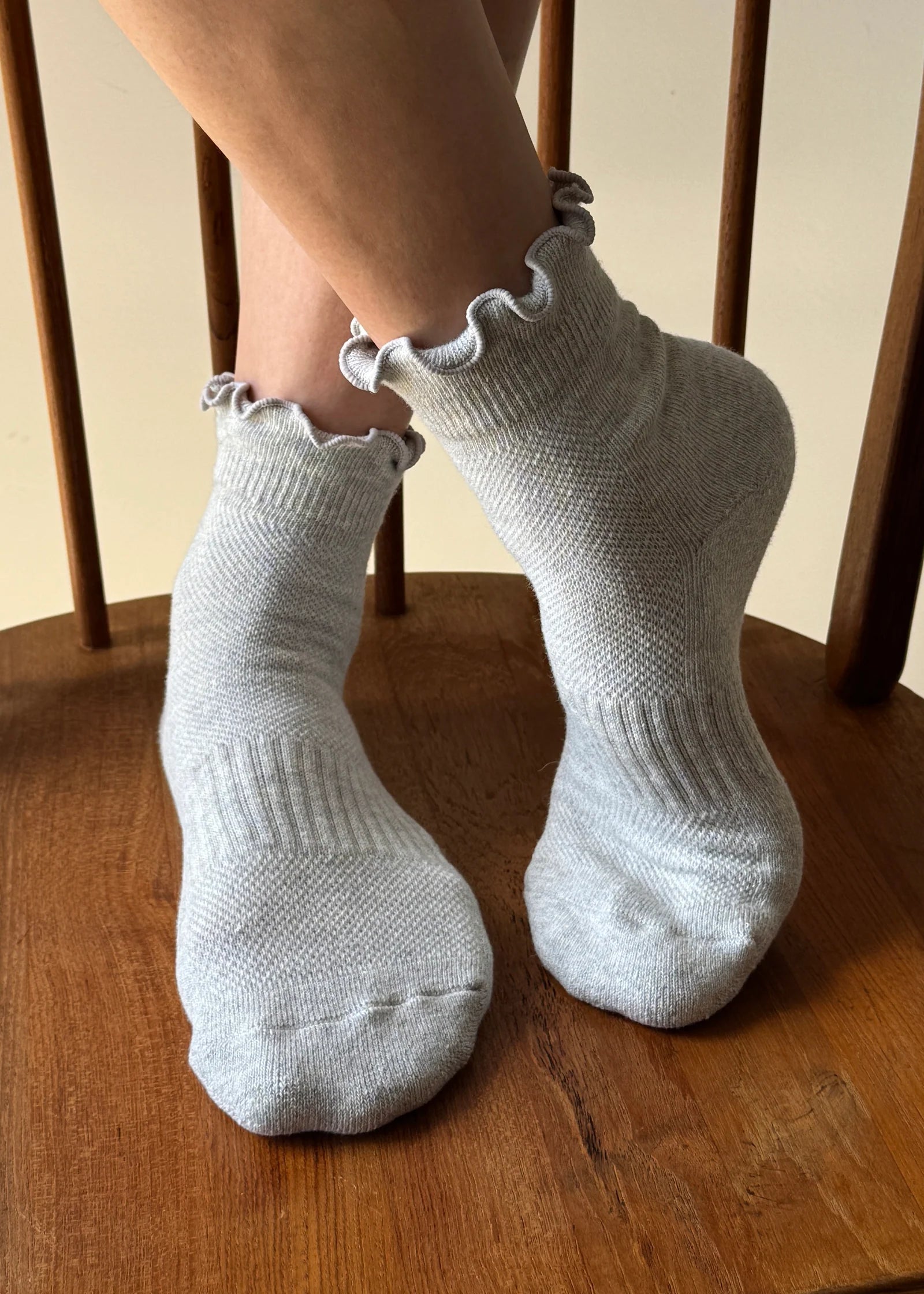 A pair of light gray ribbed ankle socks with ruffled edges worn on feet resting on a wooden chair.