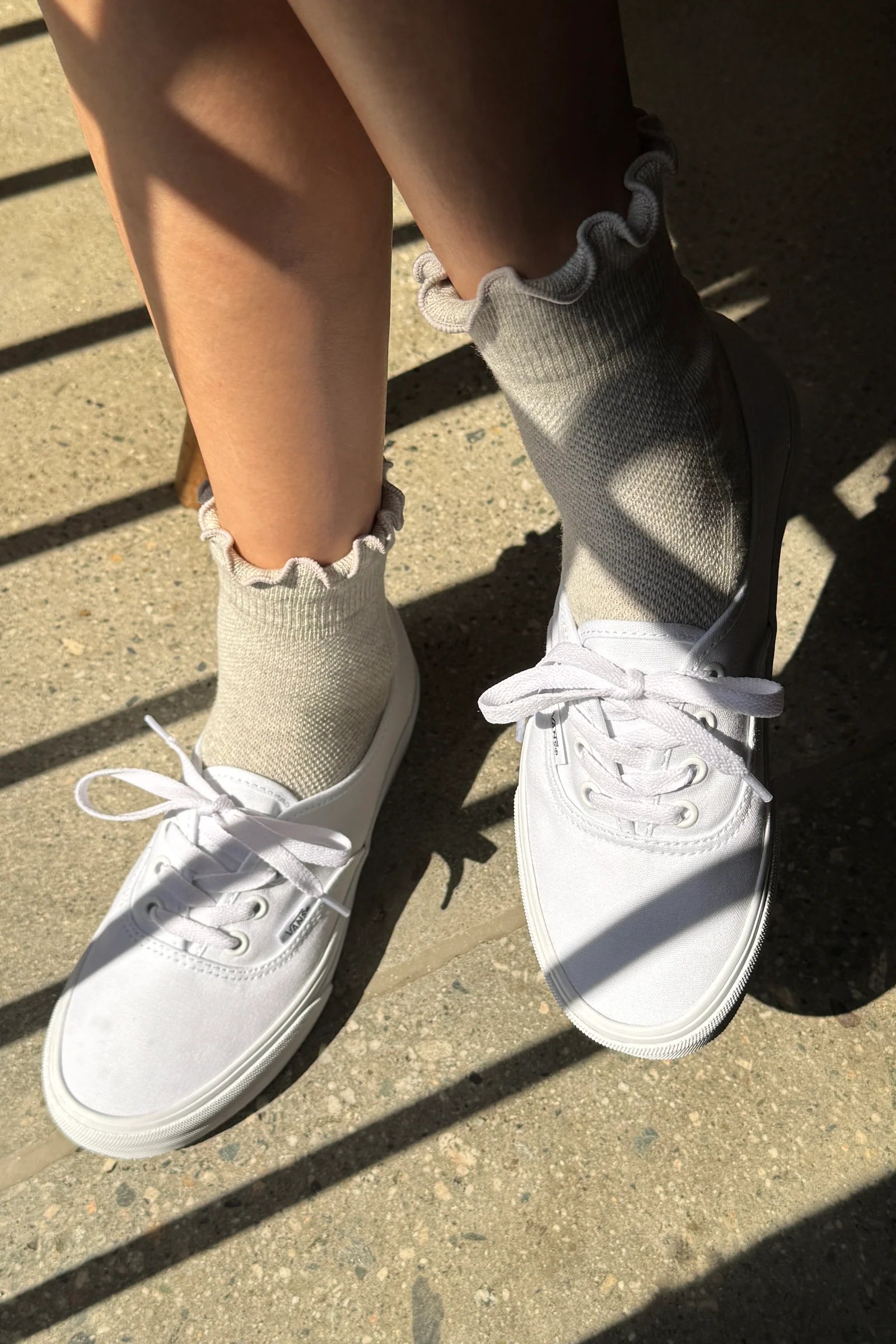 White canvas low-top sneakers worn with light beige ruffled ankle socks, shown outdoors on concrete with strong sunlight and shadows.