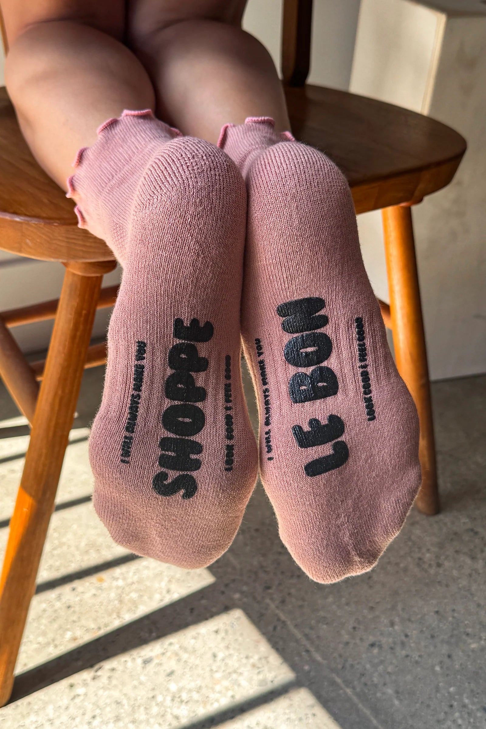 A person sitting on a wooden chair with their legs raised, wearing dusty pink ankle socks with black printed text reading 'SHOPPE' and 'LE BON'.