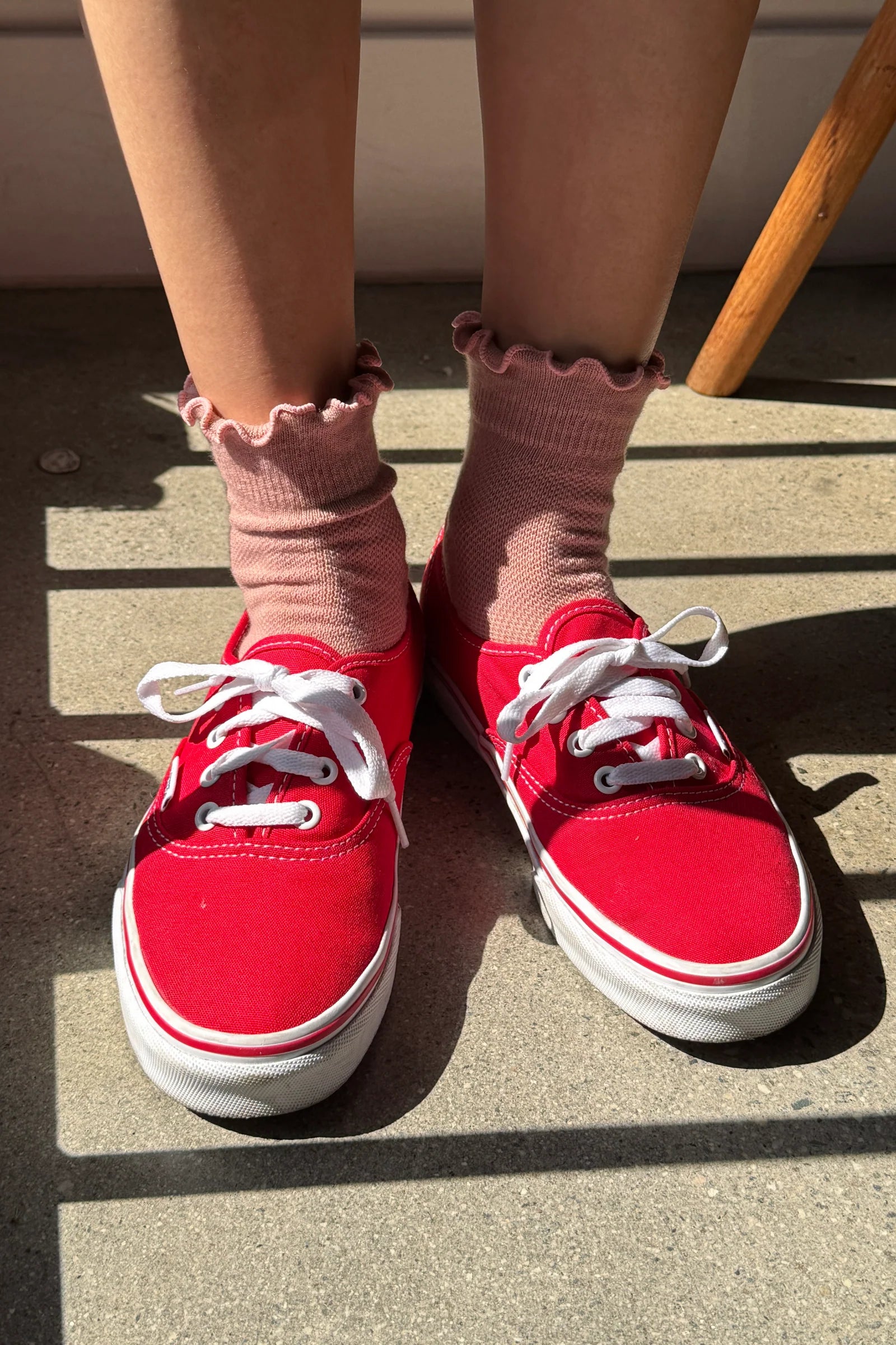 Red canvas low-top sneakers with white laces and white rubber soles, worn with pink ruffled ankle socks on a concrete floor.