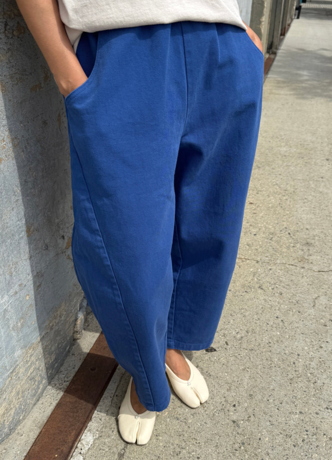 A person wearing wide-leg blue pants with a light gray top and off-white tabi-style shoes, standing outdoors against a concrete wall.