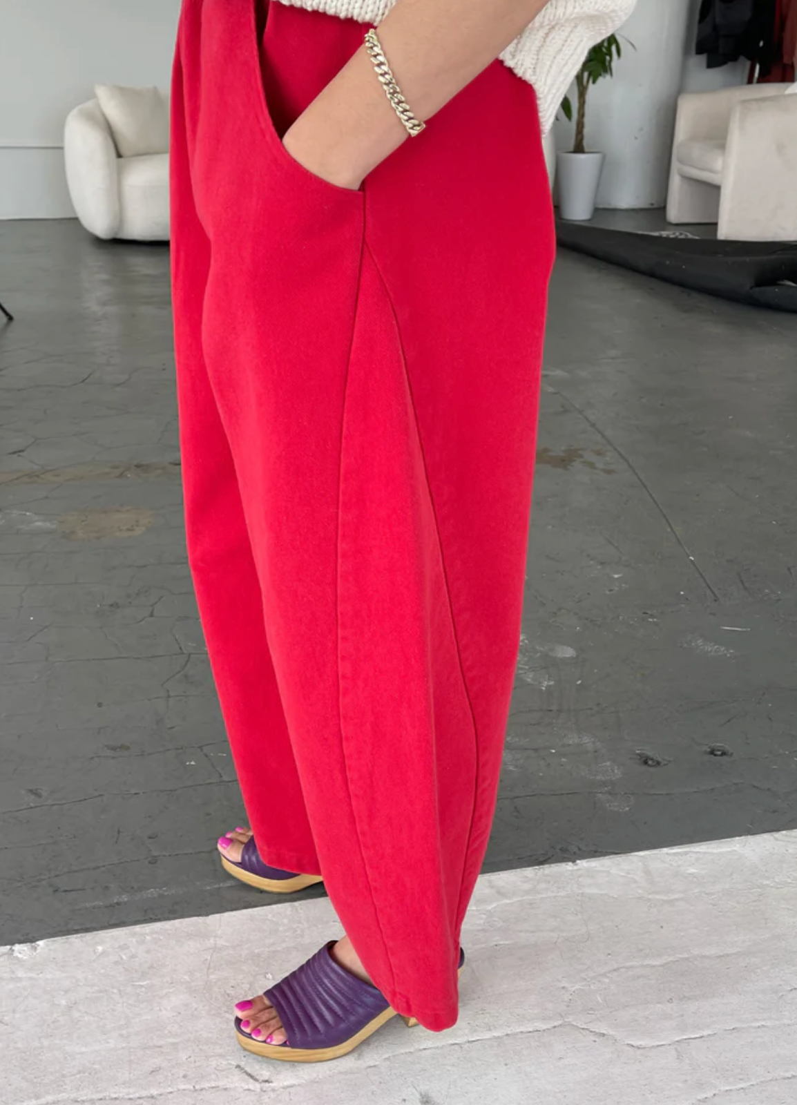 A person wearing bright red wide-leg pants with side pockets, paired with a white knit top and purple slide sandals, standing in a modern indoor studio space.