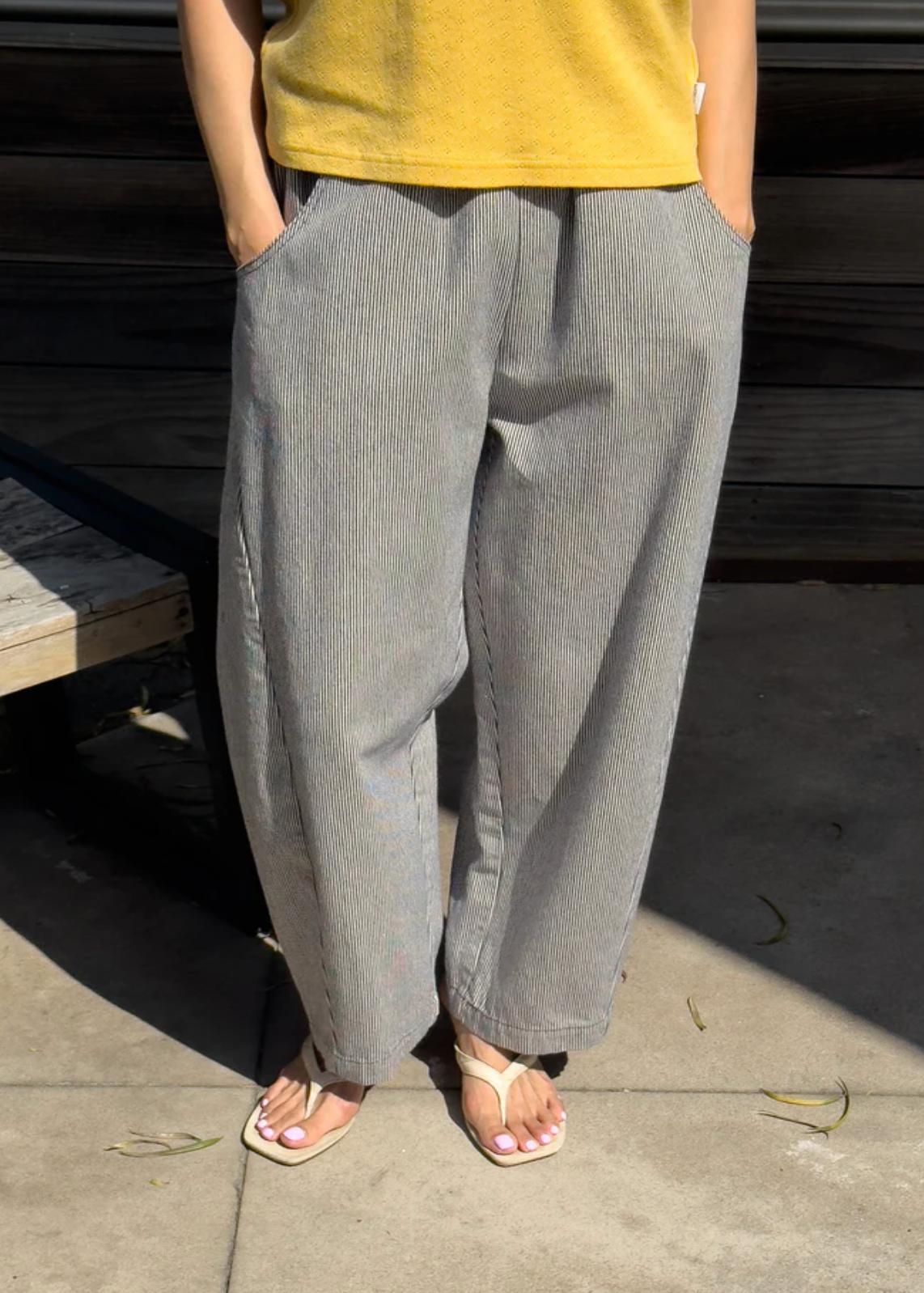 A person wearing loose, wide-leg  striped pants with a yellow top and light-colored sandals, standing outdoors on a concrete surface with a dark wooden wall behind them.