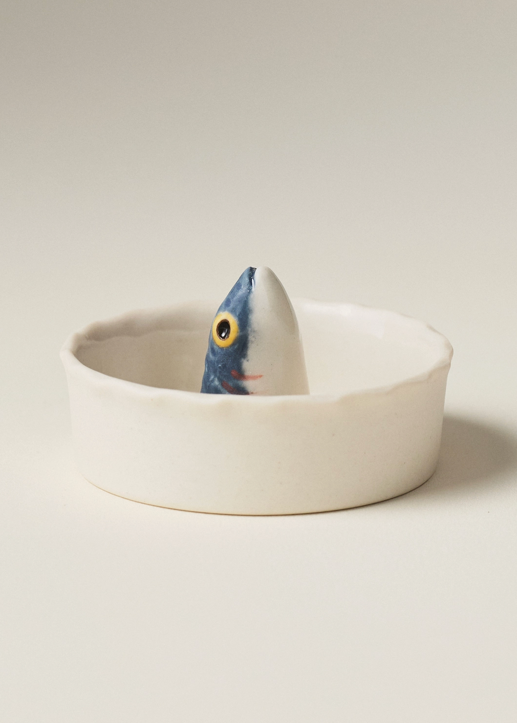 A white ceramic dish with a small painted fish figurine peeking out from the center, set against a plain light beige background.