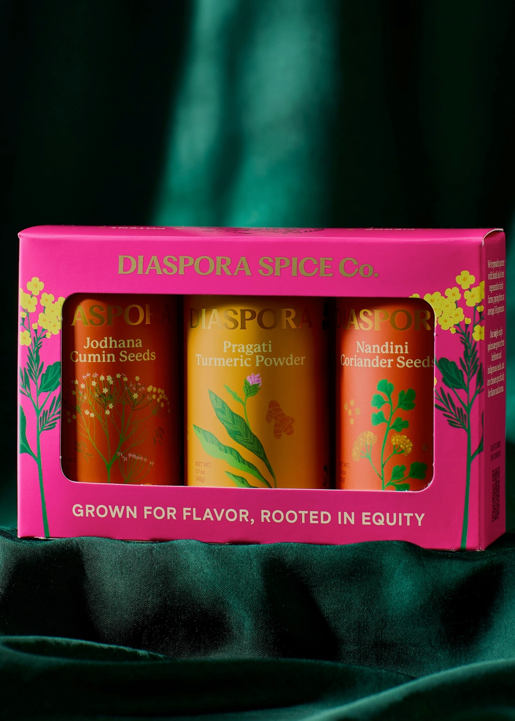 A boxed spice set from Diaspora Spice Co. in a bright pink box with three visible spice jars inside, placed on dark green draped fabric.