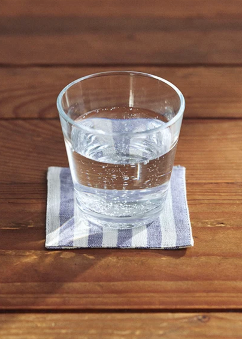 A clear glass tumbler filled with water sits on a blue-and-white striped cloth coaster on a brown wooden tabletop.