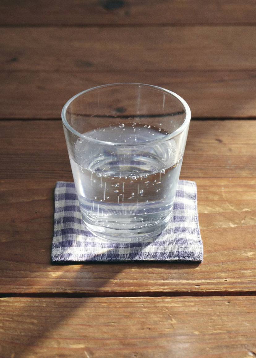 A clear glass tumbler filled with water sits on a small blue-and-white checkered cloth coaster on a rustic wooden tabletop.