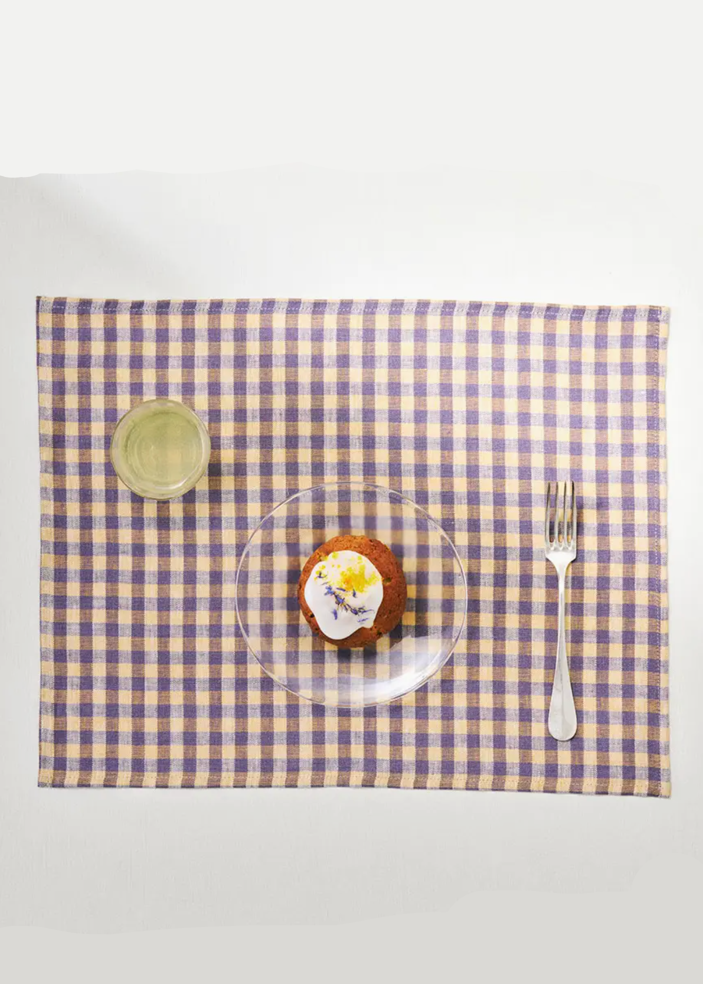 A rectangular gingham placemat in lavender and white checkered pattern, styled with a small plate of dessert, a fork, and a small dish on a white background.