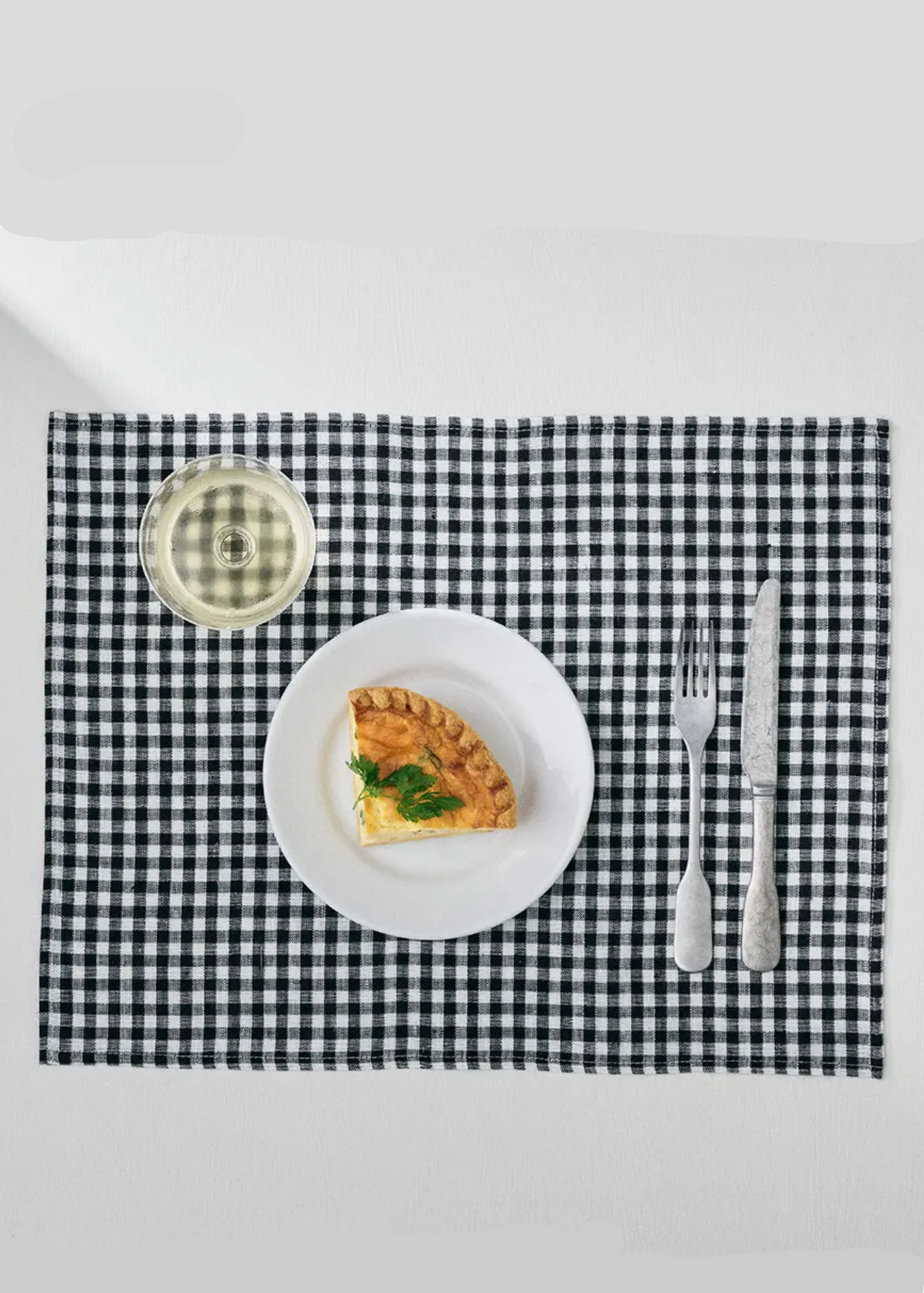 A black and white gingham placemat styled on a white table with a white plate, a slice of quiche, a fork, and a knife.