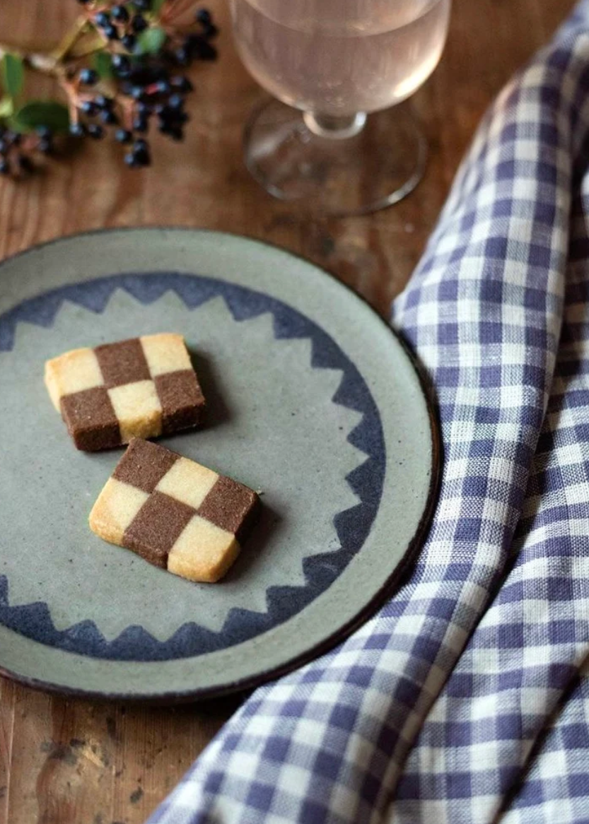 Two square checkered cookies on a gray ceramic plate with a blue patterned rim, set on a wooden table next to a plum-and-white checkered cloth and a glass of white wine.