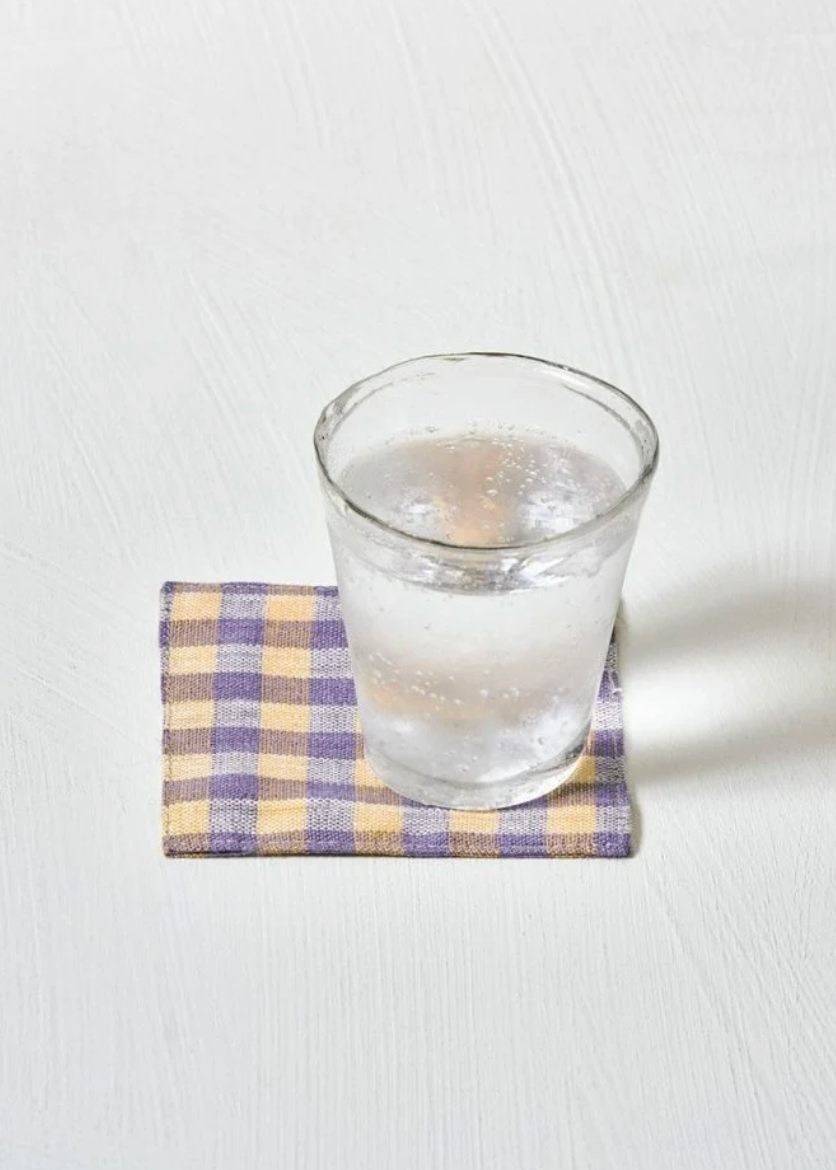 A clear glass tumbler filled with water sits on a small checkered napkin in yellow, purple, and white, placed on a light textured white surface.