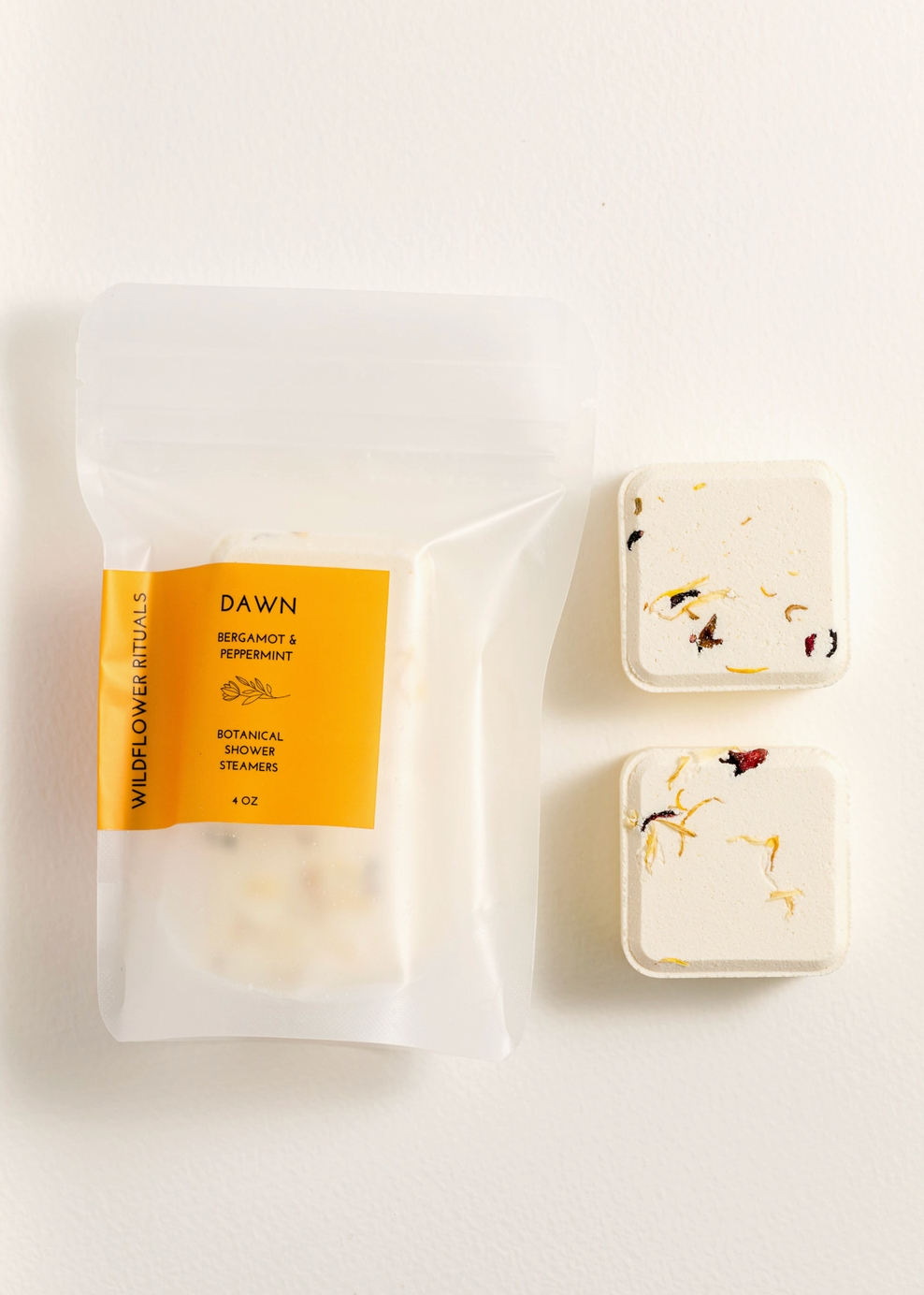A clear resealable pouch labeled DAWN Botanical Shower Steamers with a yellow-orange label, shown next to two small square cream-colored bath steamers with dried botanical inclusions, on a clean white background.