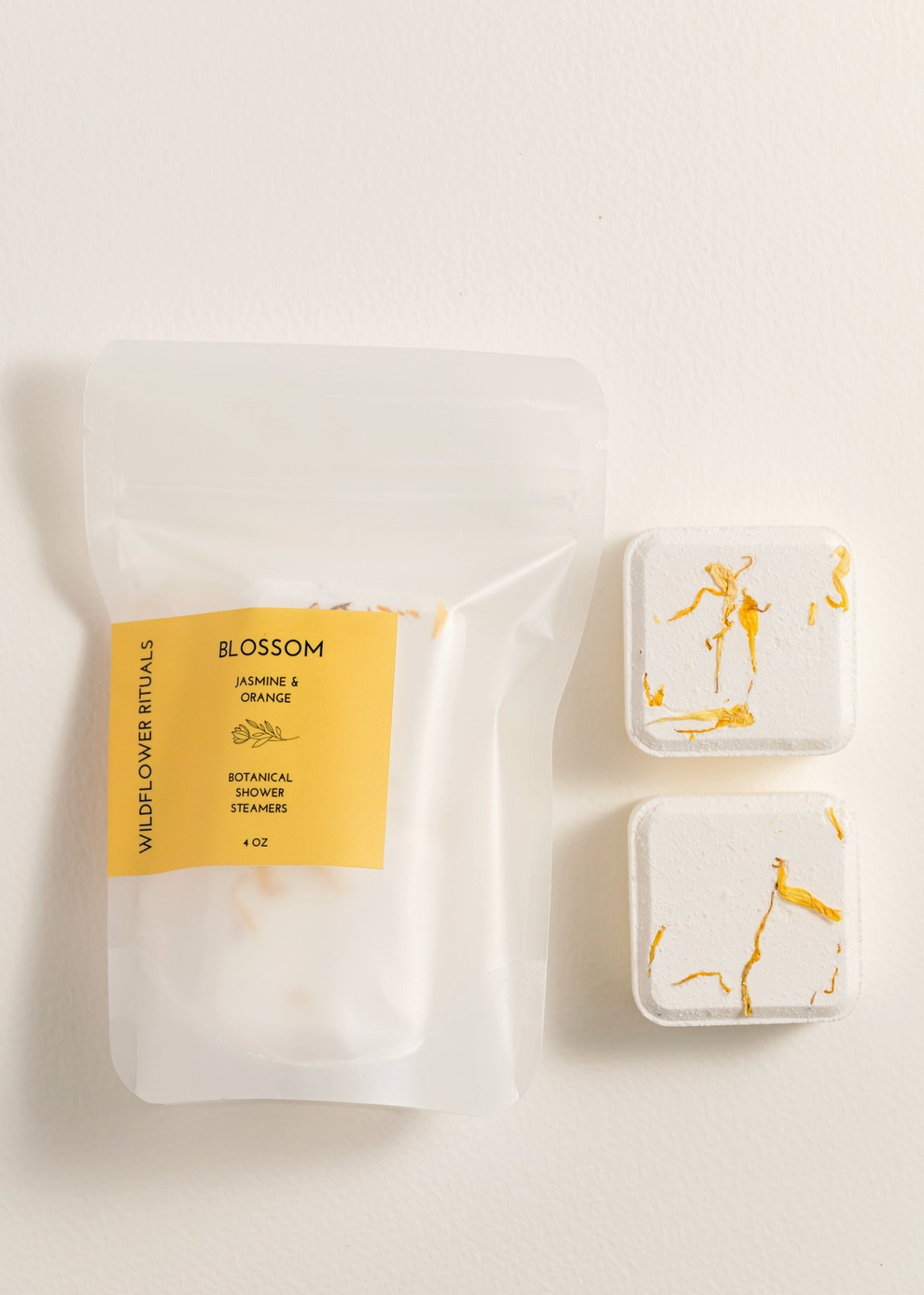 A clear stand-up pouch labeled BLOSSOM Jasmine & Orange by Wild Flower Rituals, shown next to two small square white bath steamers