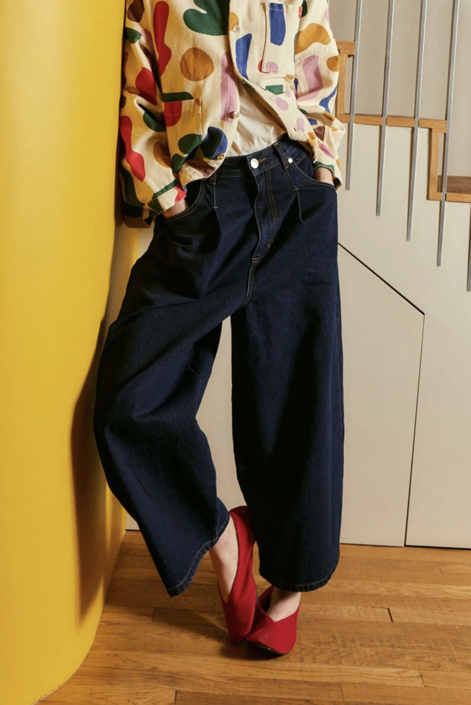 person wearing wide leg jeans with colorful jacket, and red shoes against a yellow wall in a living setting