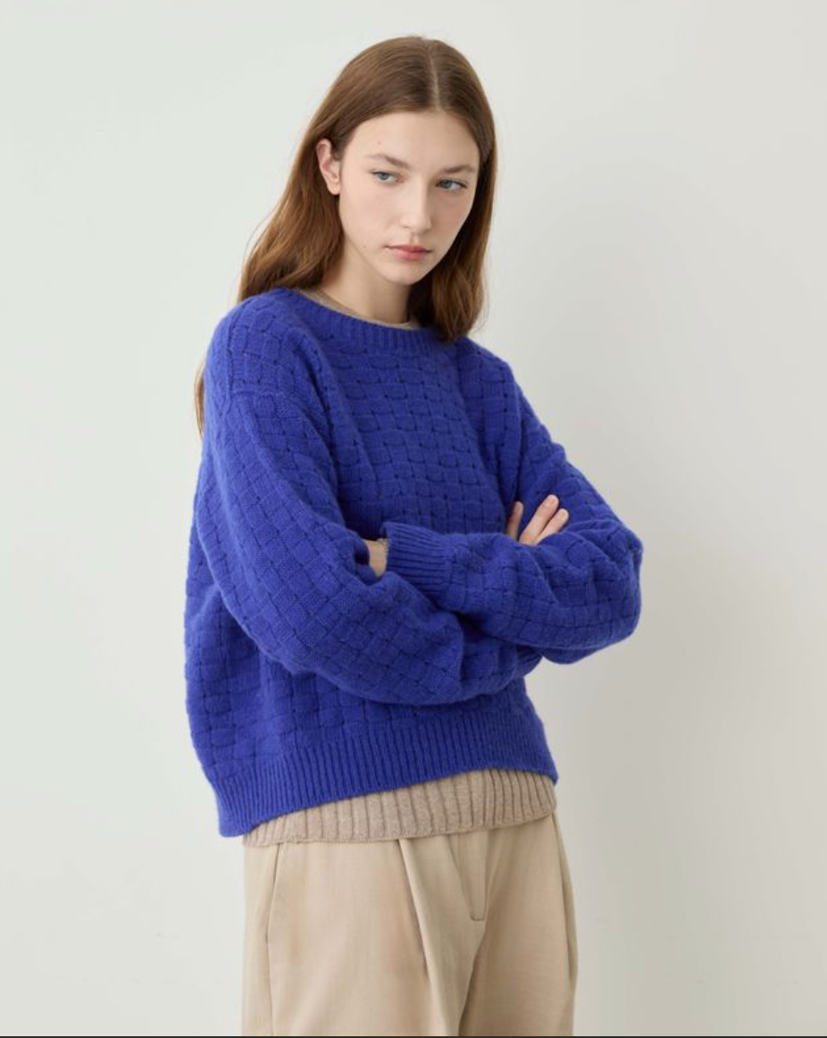A woman wearing a vibrant royal blue chunky knit sweater with a textured diamond pattern, paired with beige trousers, standing against a plain off-white background.