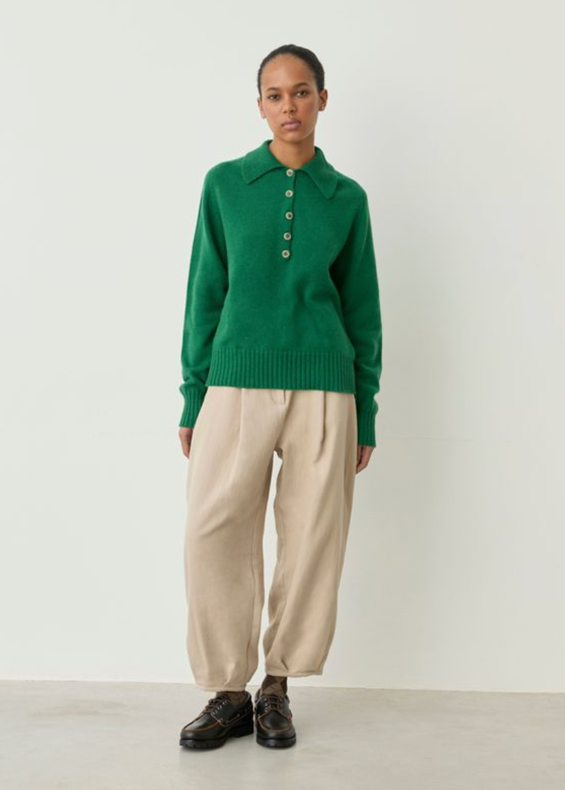 A model stands against a plain off-white studio wall wearing a green knit polo sweater with gold buttons, beige wide-leg trousers, and dark brown leather shoes.