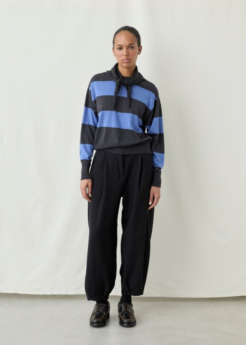 A model stands against a plain off-white studio backdrop wearing a cropped knit hoodie with wide horizontal stripes in dark charcoal gray and medium blue, paired with loose black pants and dark brown shoes.