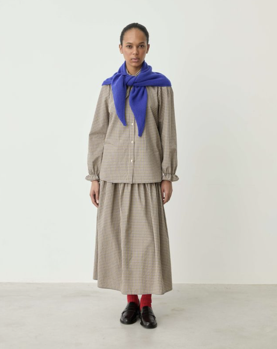 A model stands against a plain off-white studio wall wearing a beige and brown gingham button-up shirt and matching skirt set, with a bright blue scarf tied around the neck, red socks, and dark brown shoes.