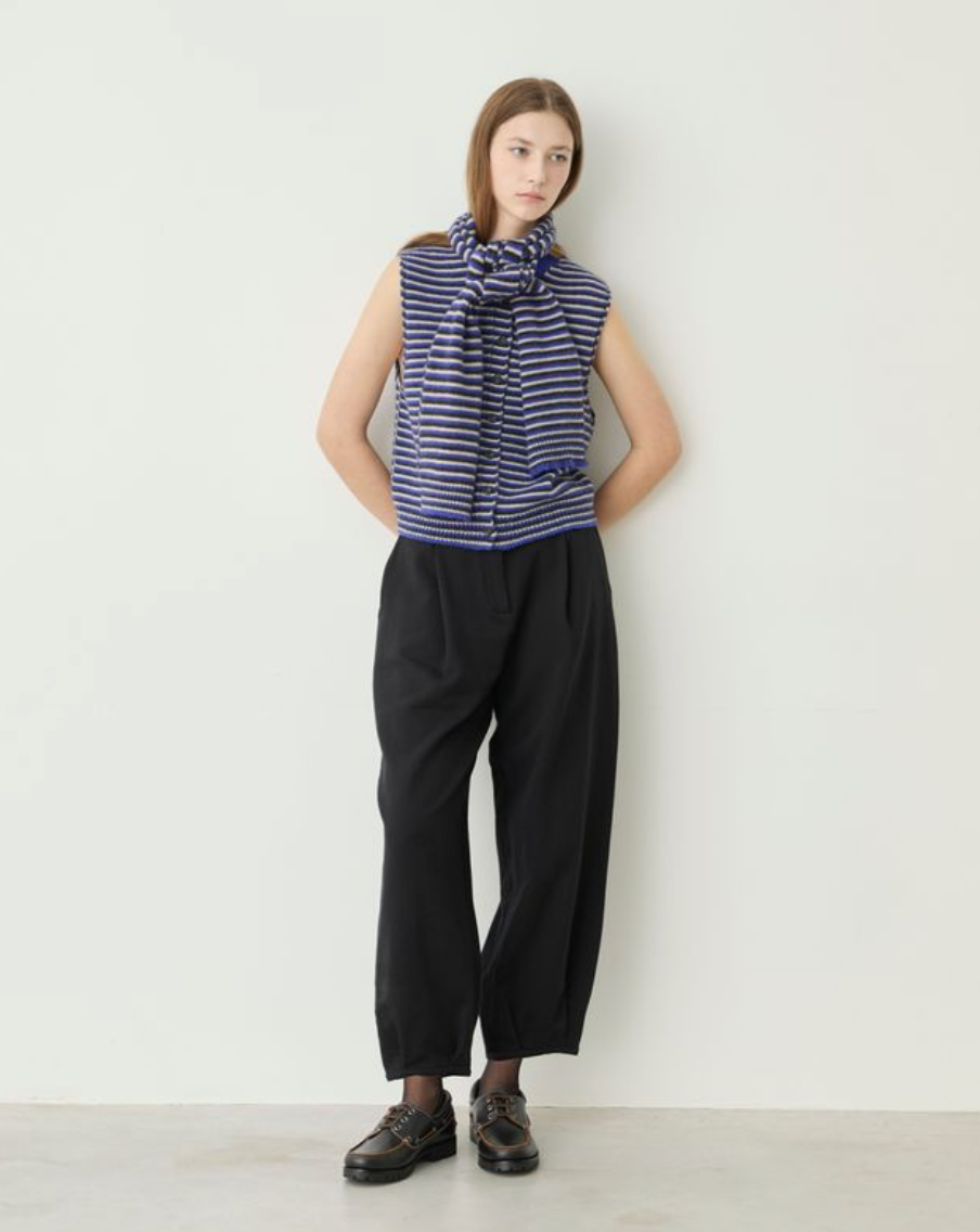 A model stands against a plain off-white wall wearing a sleeveless navy and white striped top with a large front bow, black wide-leg cropped pants, and dark brown leather shoes.