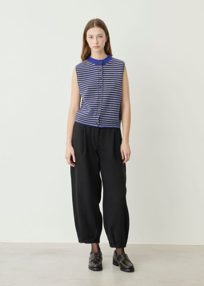 A model stands against a plain off-white studio wall wearing a sleeveless knit top with blue and navy horizontal stripes, black wide-leg cropped pants, and black leather shoes.