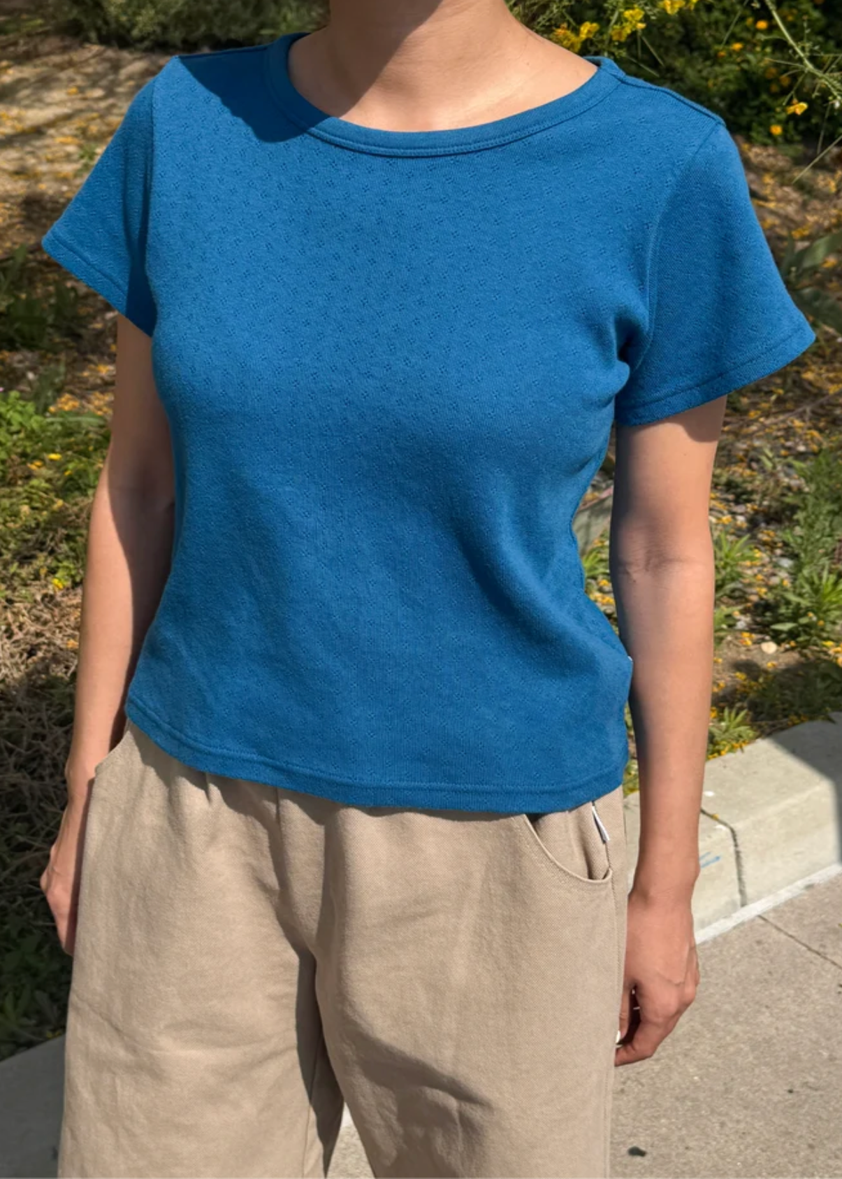A person wearing a short-sleeve crew-neck t-shirt in a heathered blue color, paired with beige linen pants, standing outdoors on a sunny day.