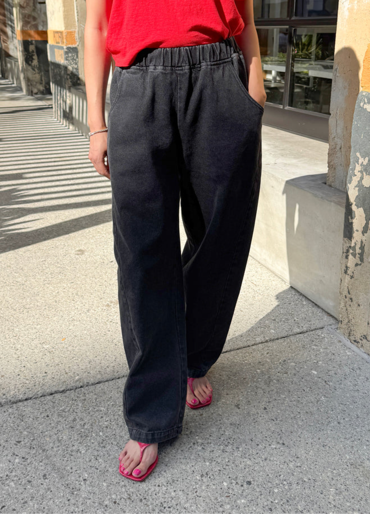 A person wearing loose black wide-leg jeans, a red top, and pink thong sandals walking on a concrete sidewalk outdoors.