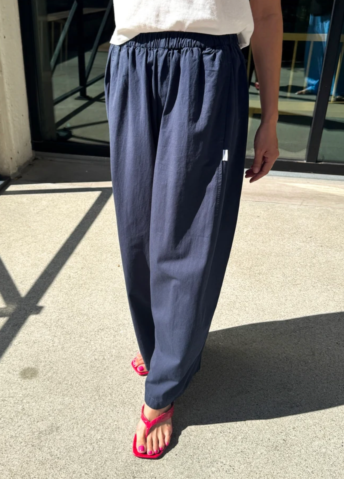 A person wearing loose navy blue drawstring pants and bright pink thong sandals, standing outdoors on a concrete surface with glass doors in the background.