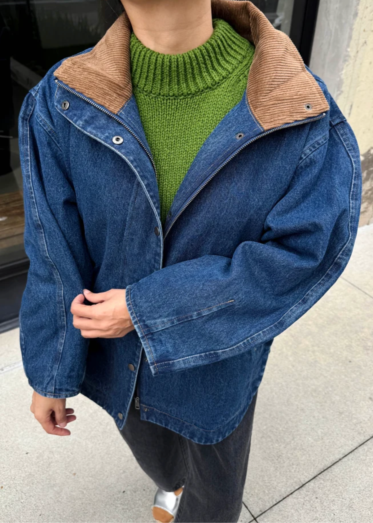 A person wearing an oversized blue denim jacket with a brown corduroy collar, layered over a green knit sweater and dark pants, standing outdoors on a concrete surface.