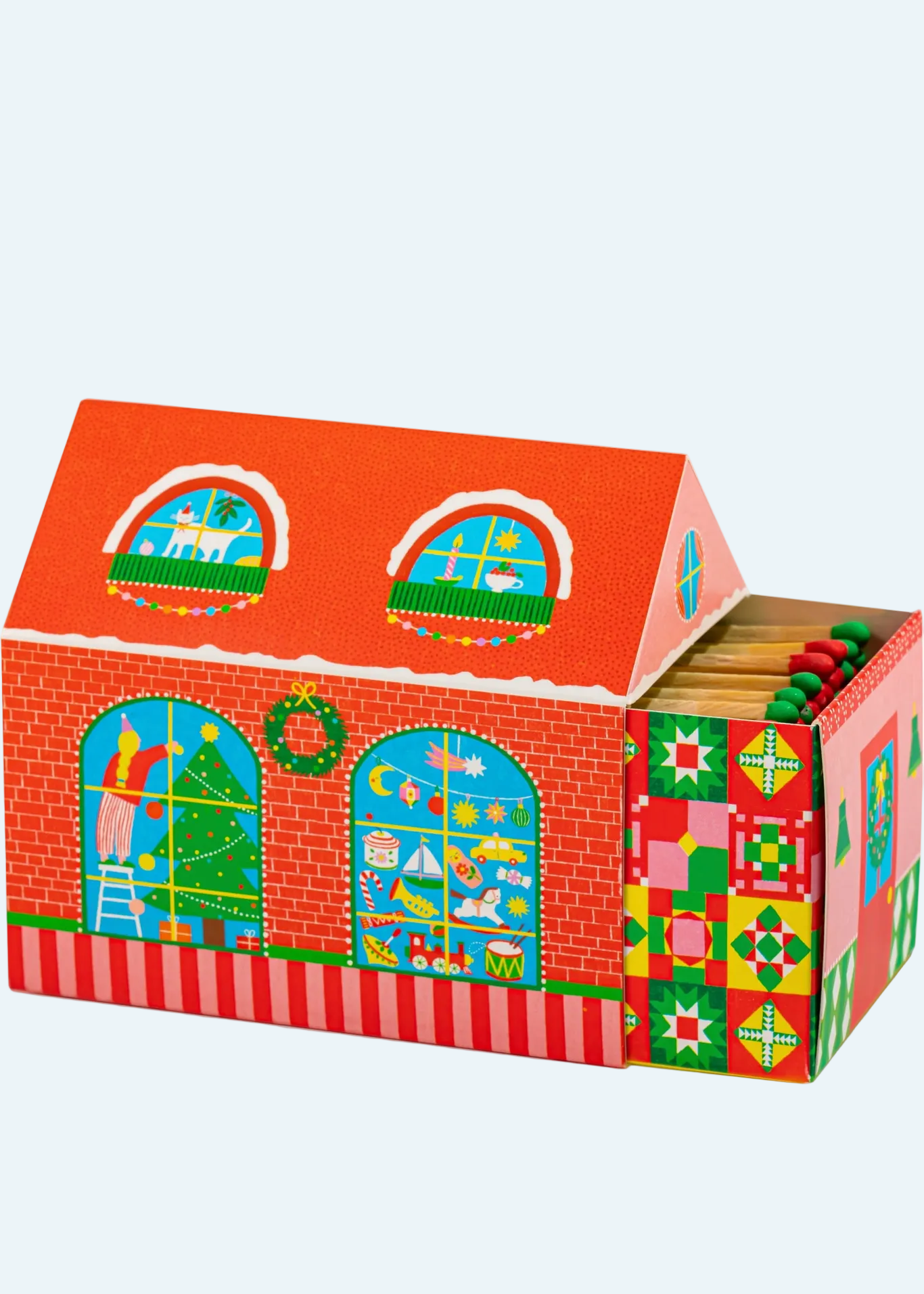 A Christmas-themed wooden matchbox box shaped like a gingerbread house, shown open to reveal wooden matches inside. The box features red brick detailing, a red-orange roof with arched windows, green wreaths, and colorful holiday patterns on the side panel.