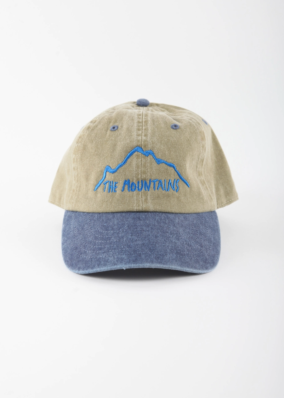 A two-tone baseball cap with a beige washed cotton crown and a blue denim-look curved brim, featuring a blue embroidered mountain and 'THE MOUNTAINS' text on the front.