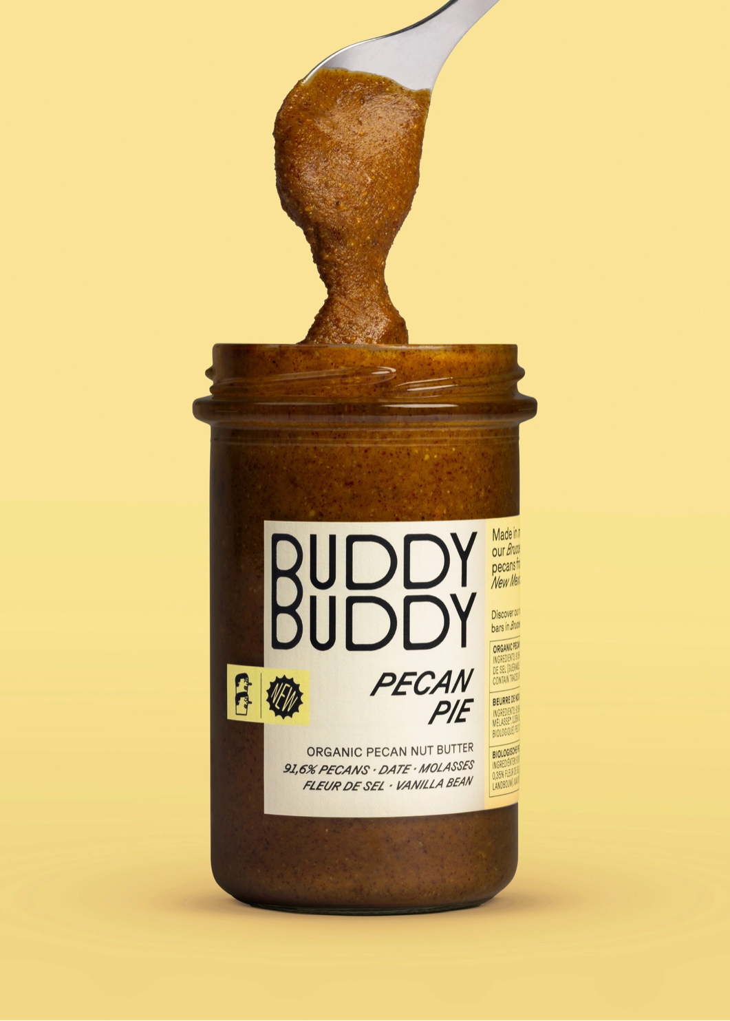 A jar of Buddy Buddy Pecan Pie nut butter with a spoon lifting the thick brown spread above the label, set against a solid pale yellow background.