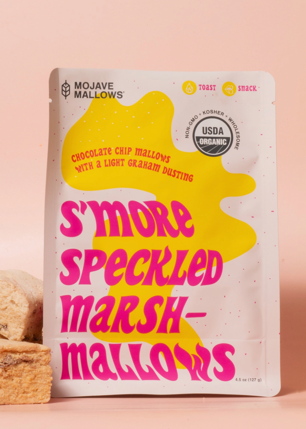 A stand-up pouch of Mojave Marshmallows Chocolate Chip Marshmallows with a pink and yellow design, shown on a peach-pink background with a few pieces of the marshmallows stacked at the lower left.