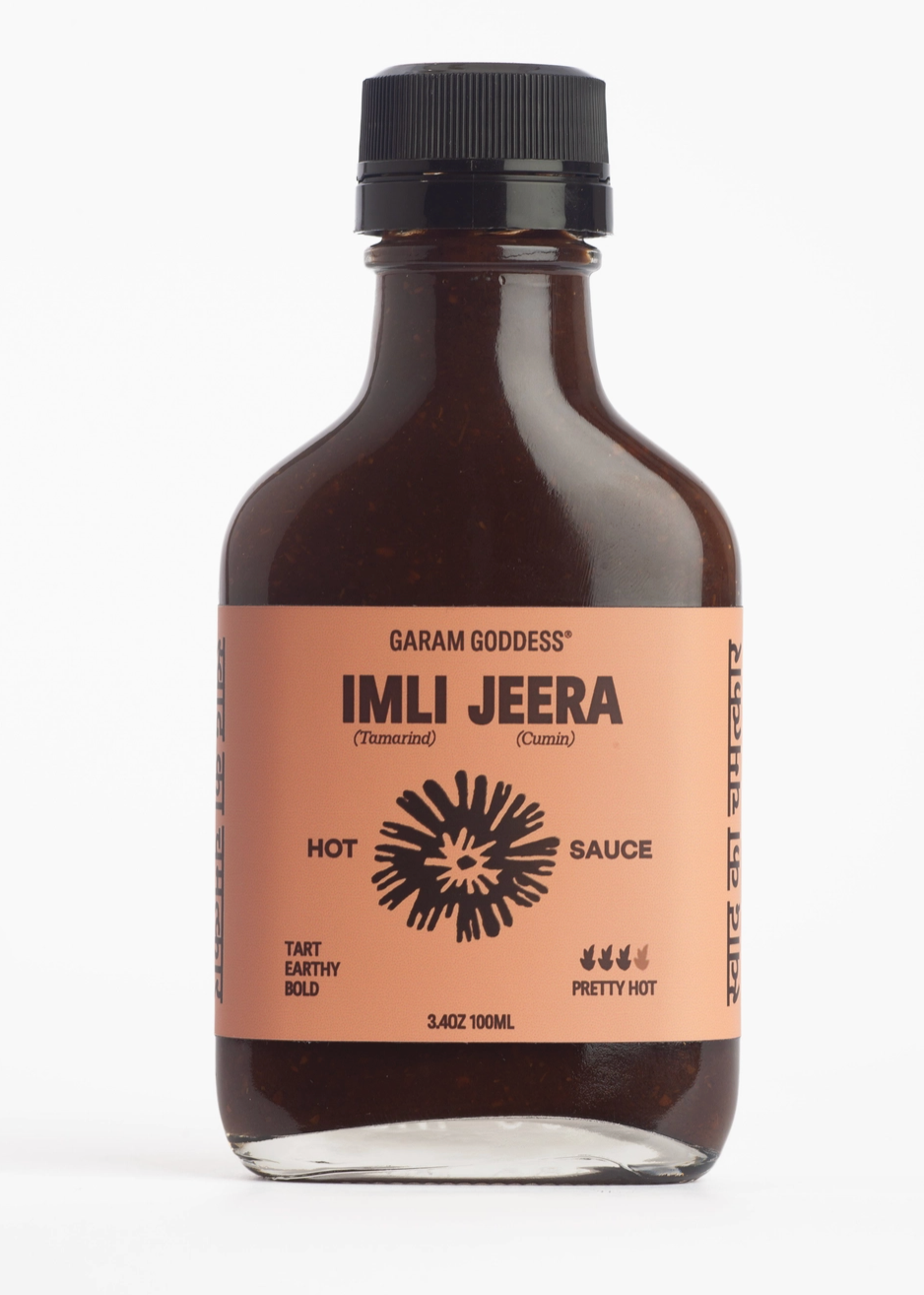 A 3.4oz glass bottle of Garam Goddess Imli Jeera Hot Sauce, featuring a black cap and a peach-colored label with black text and a starburst graphic, set against a plain white background.