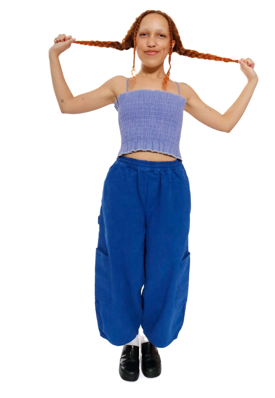 A woman with long red braids poses against a white background, wearing a light purple ribbed crop top and bright blue wide-leg pants.