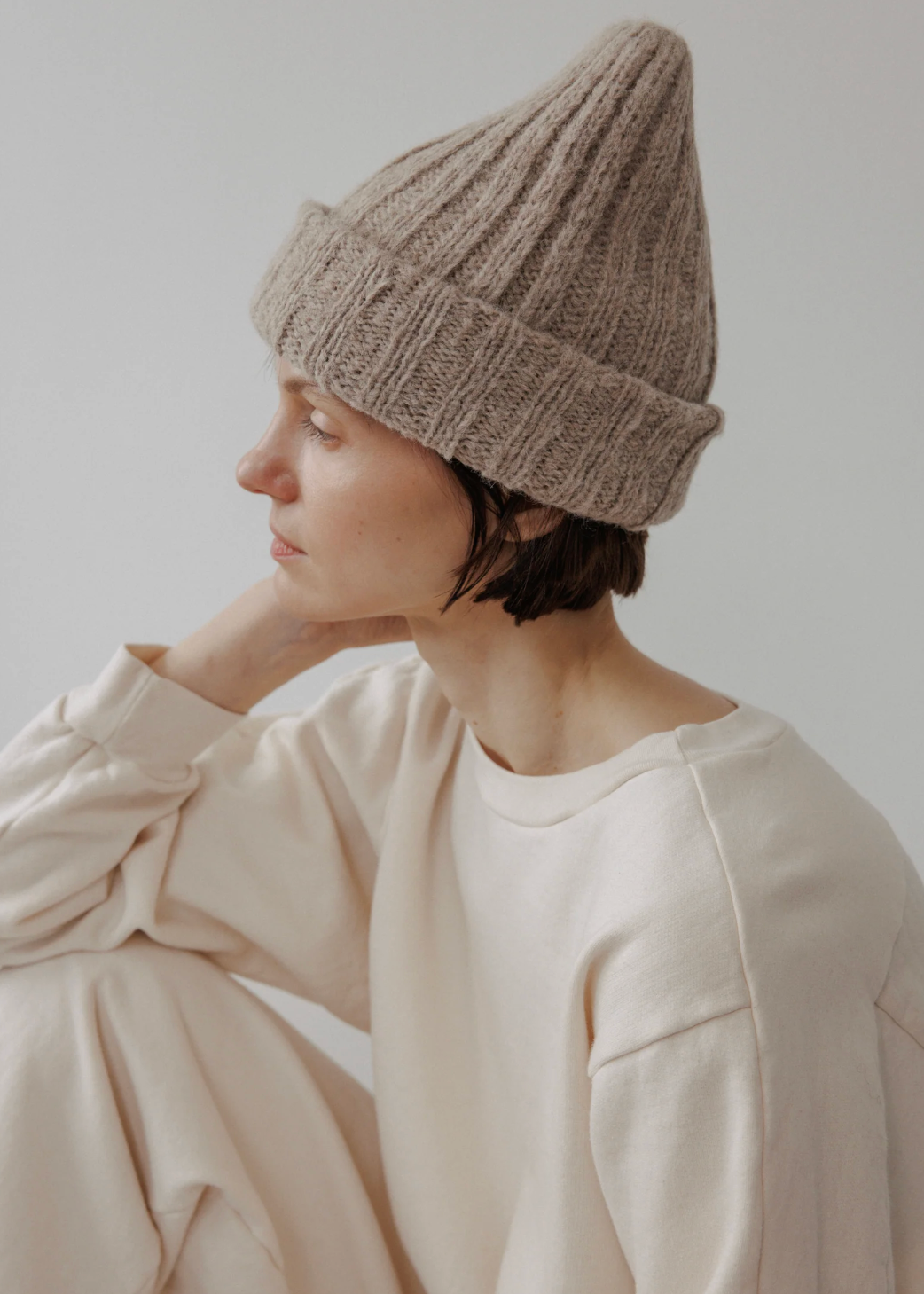 A person wearing a beige ribbed knit beanie and a cream sweatshirt, shown in profile against a plain light gray background.