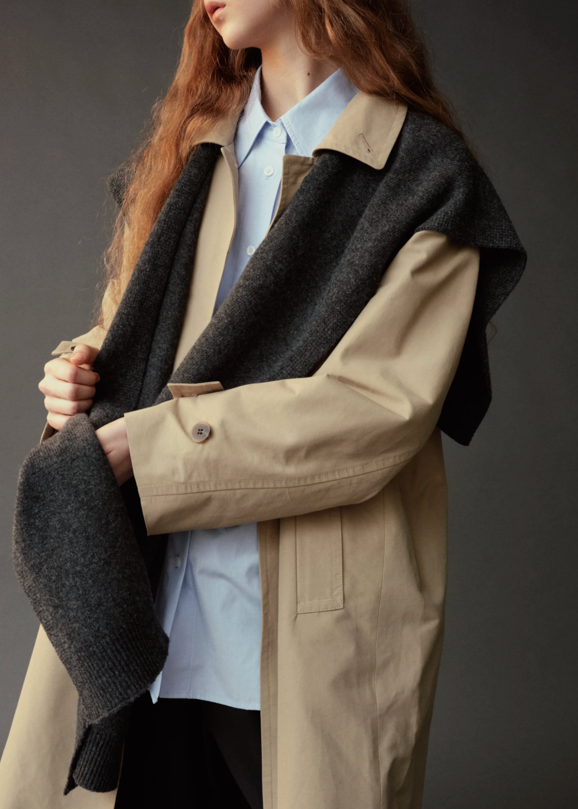 A model wearing a beige trench coat with dark gray knit shoulder panels and a light blue collared shirt, photographed against a dark gray studio background.