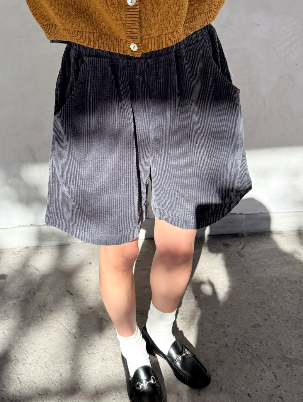 Person wearing a brown cardigan, black and white checkered skirt, white socks, and black shoes on a gray floor.