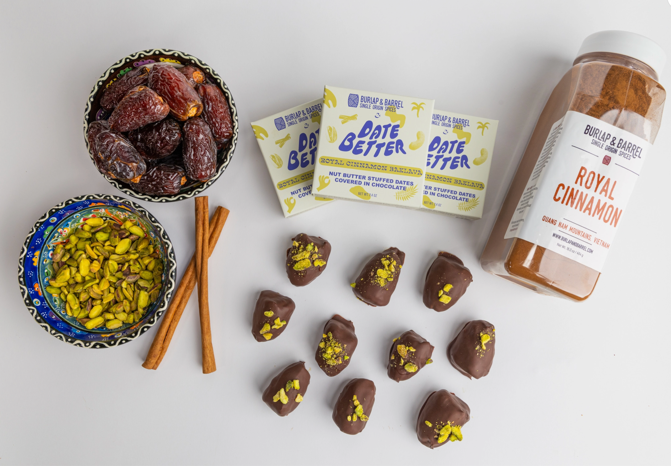 Chocolate-dipped treats with pistachios, bowls of dates and nuts, and 'Date Better' and 'Royal Cinnamon' products on a white surface.