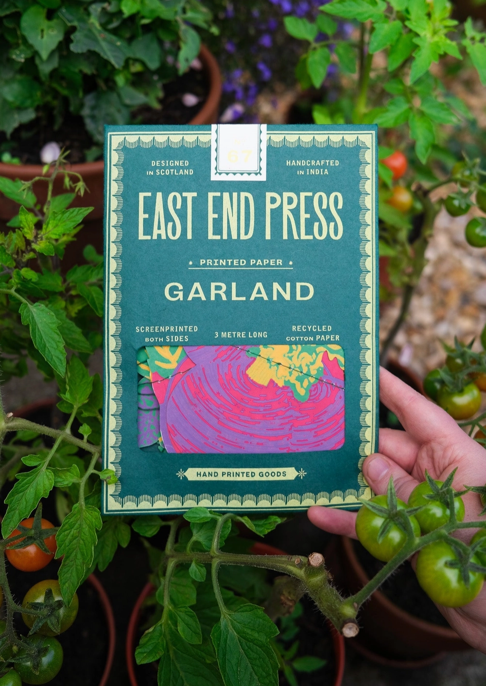 Handheld 'East End Press Garland' printed paper package with plants in the background