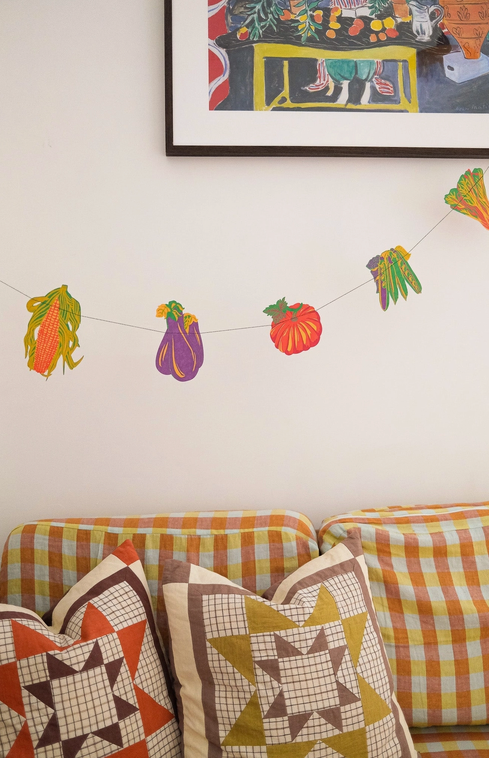 Decorative string with colorful paper vegetables hanging on a wall above a sofa with patterned cushions.