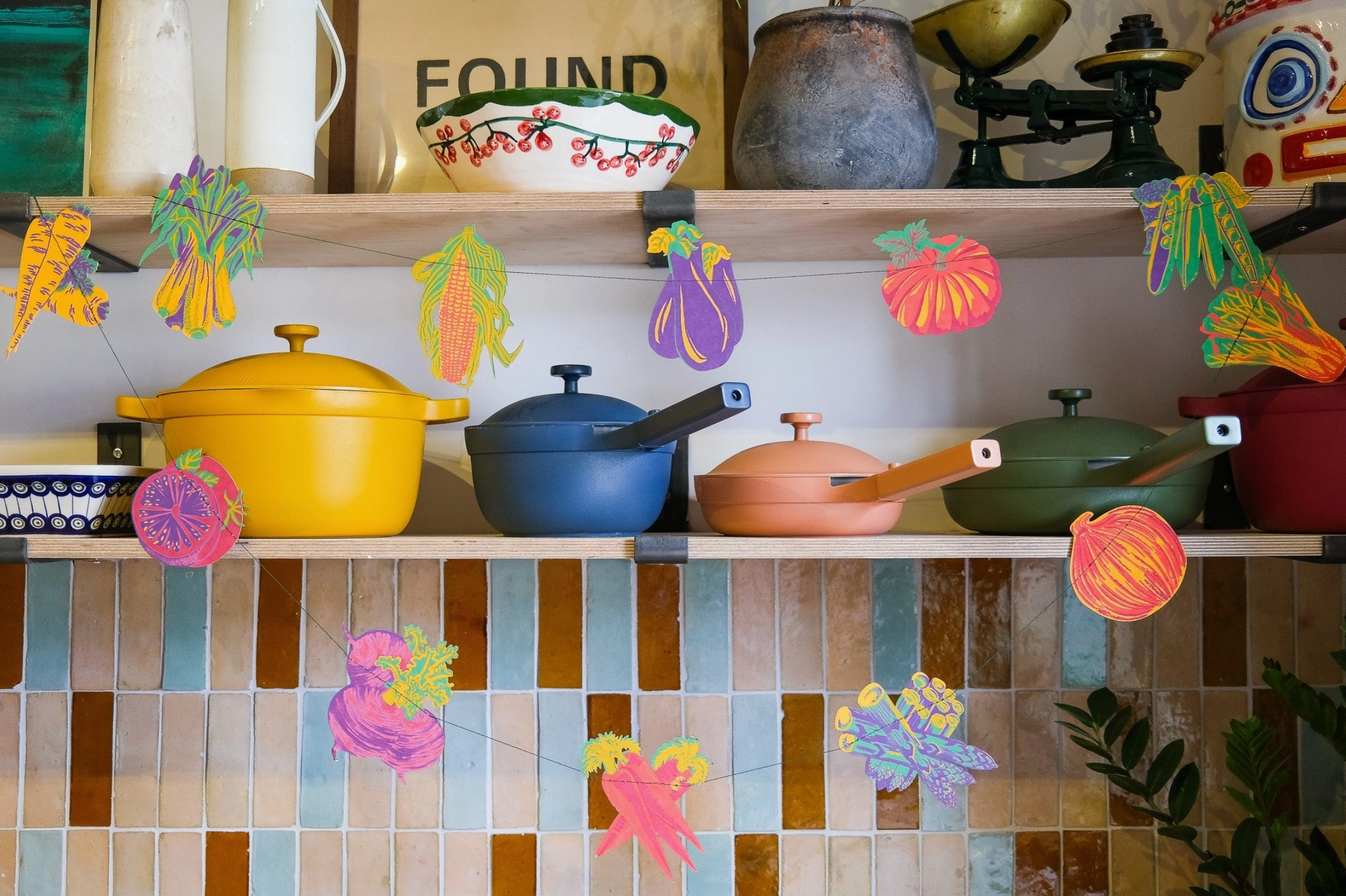 Colorful ceramic pots on a shelf with decorative cutout vegetables against a tiled wall.