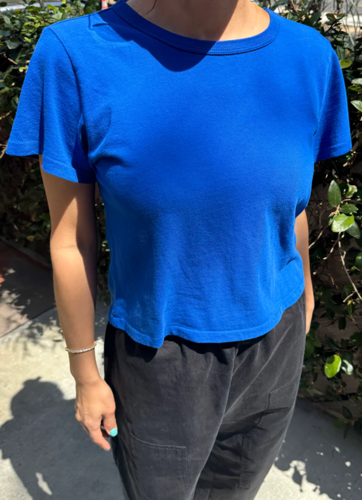 Person wearing a blue t-shirt and dark pants standing outdoors with greenery in the background