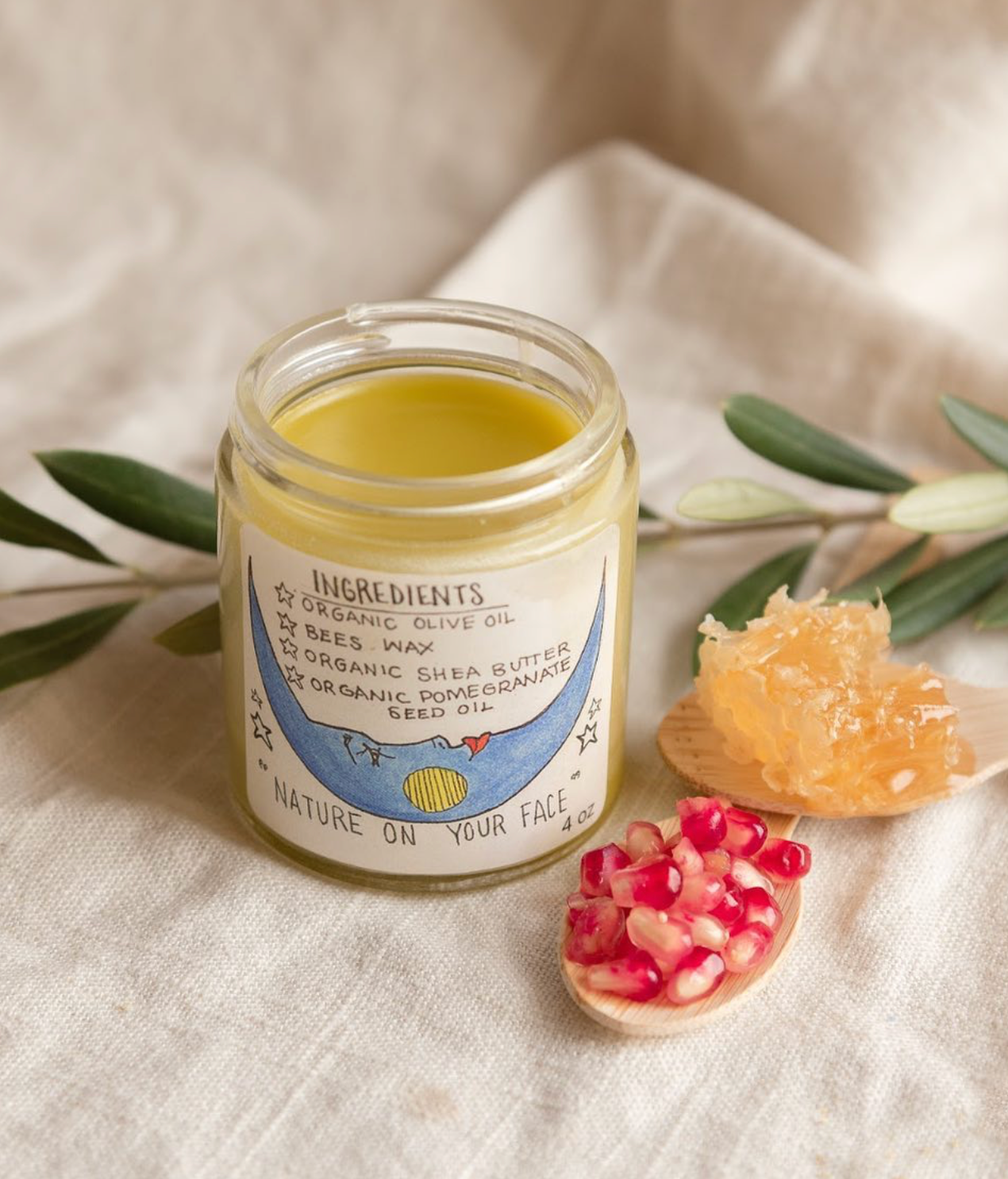 Jar of natural skincare product with ingredients listed, surrounded by honeycomb, pomegranate seeds, and olive leaves on a textured fabric background.