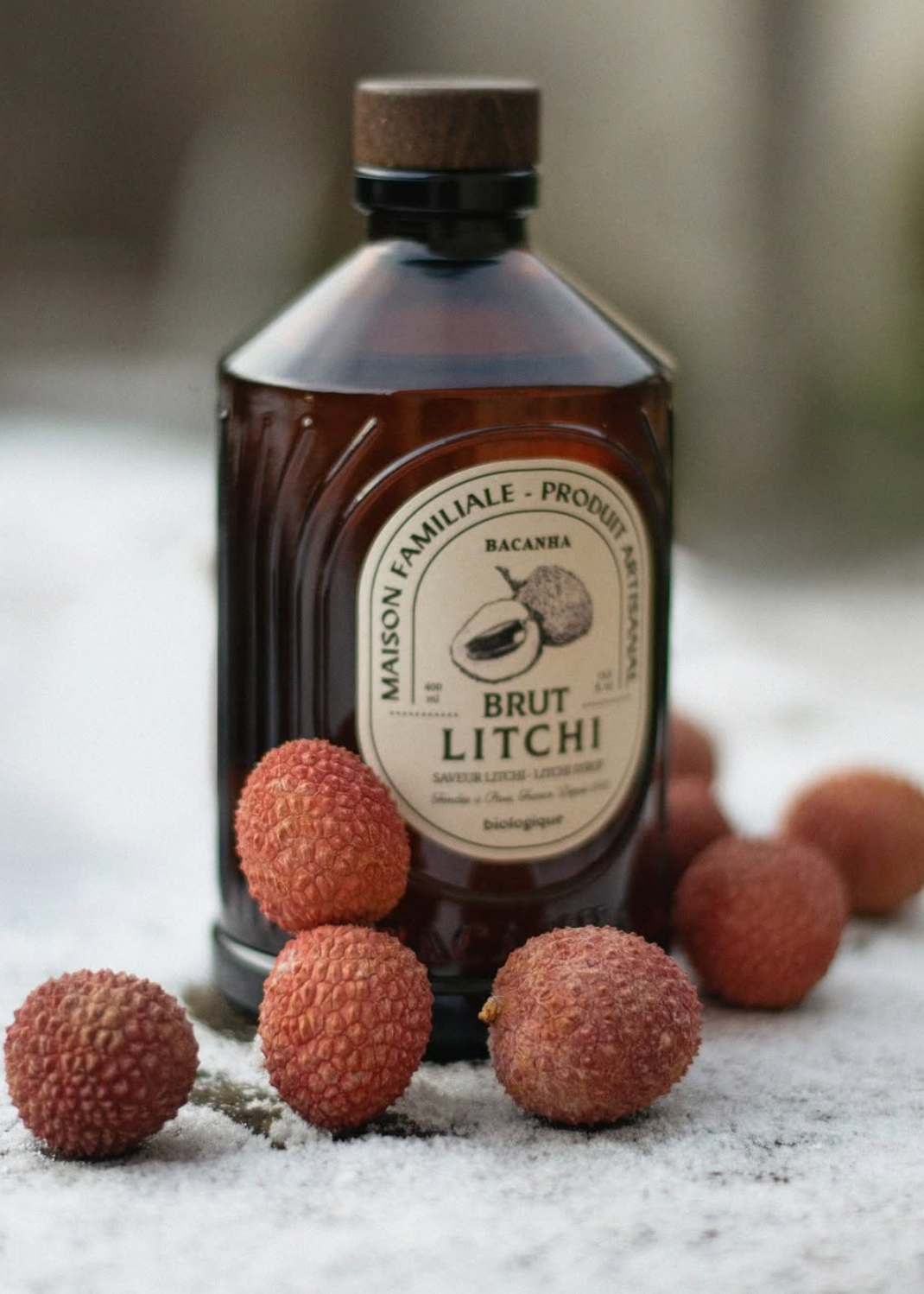 A dark amber glass bottle of Maison Familiale - Produit Brut Litchi syrup with a cork stopper, surrounded by fresh lychee fruits on a light textured surface with a soft, blurred background.