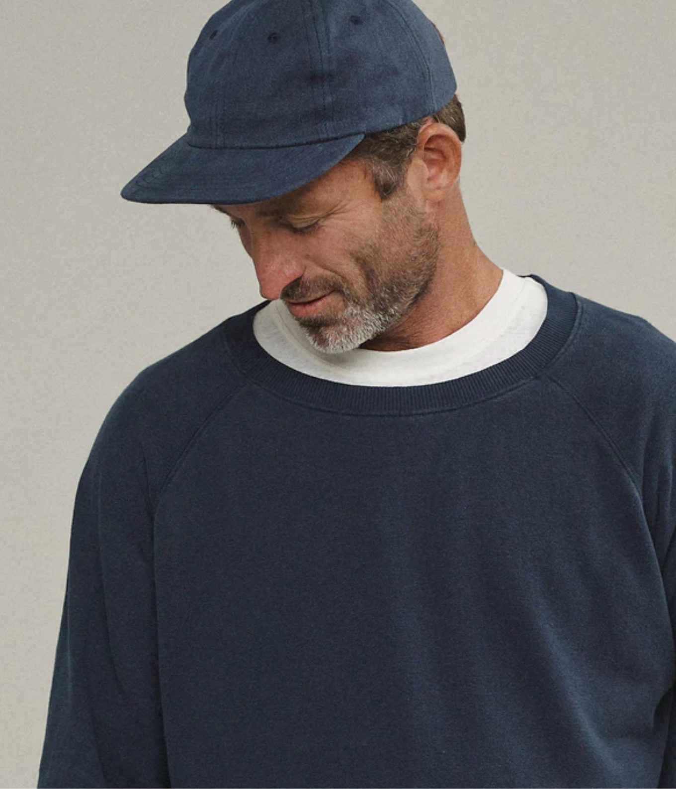 A man wearing a navy blue crewneck sweatshirt layered over a white t-shirt, paired with a matching navy blue baseball cap, set against a plain light gray background.