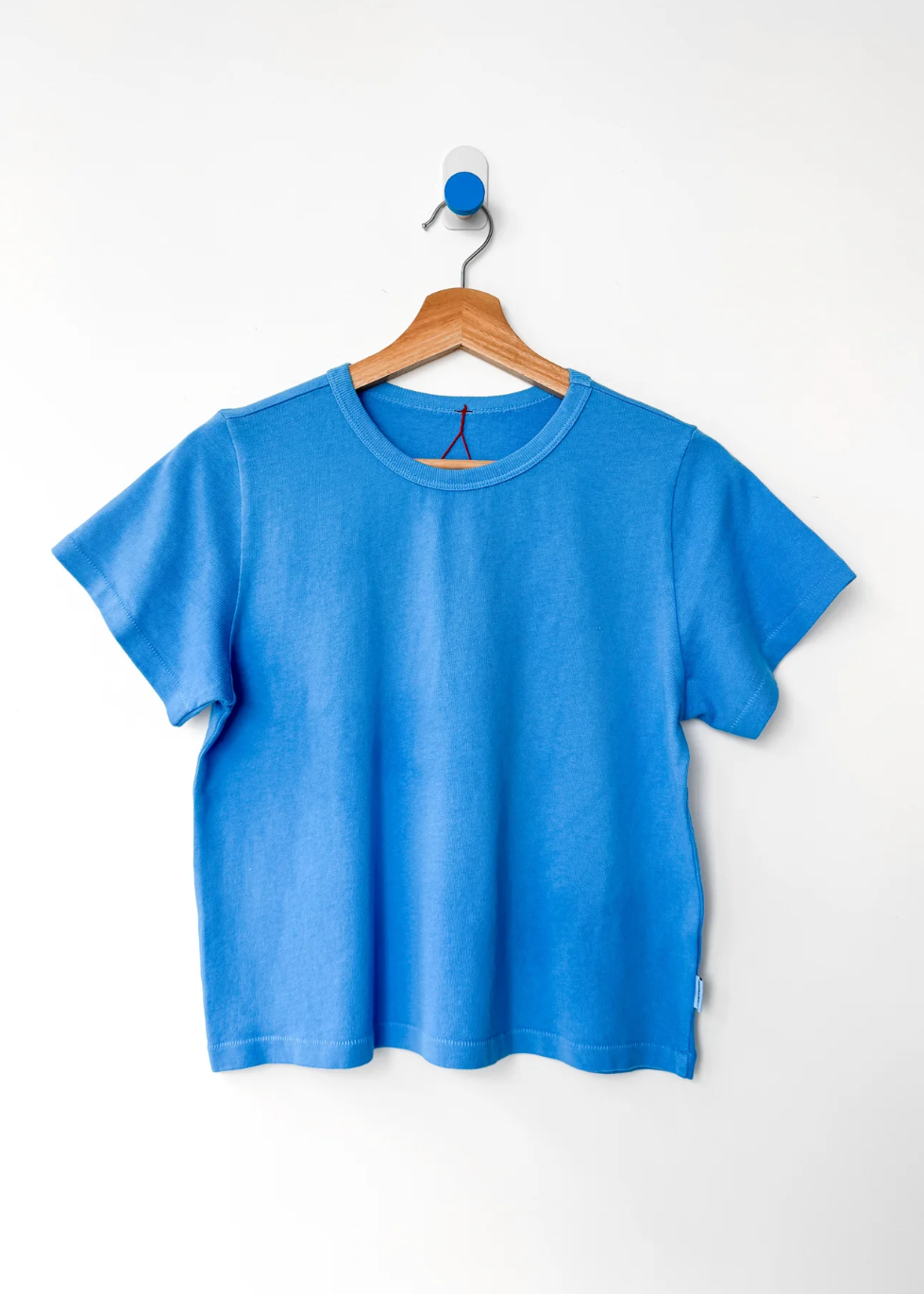 A bright blue short-sleeve crewneck t-shirt hanging on a wooden hanger from a blue hook against a plain white wall.