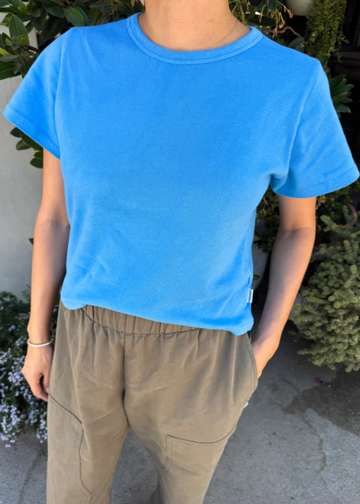 A person wearing a bright blue short-sleeve crewneck t-shirt and khaki pants, standing outdoors in front of green plants.