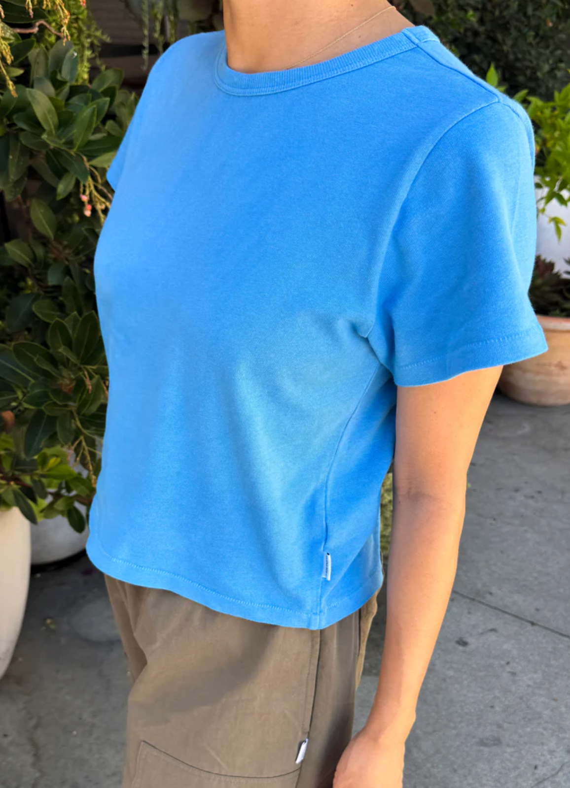 A person wearing a bright blue short-sleeve crewneck crop top and khaki pants, standing outdoors in front of green plants.