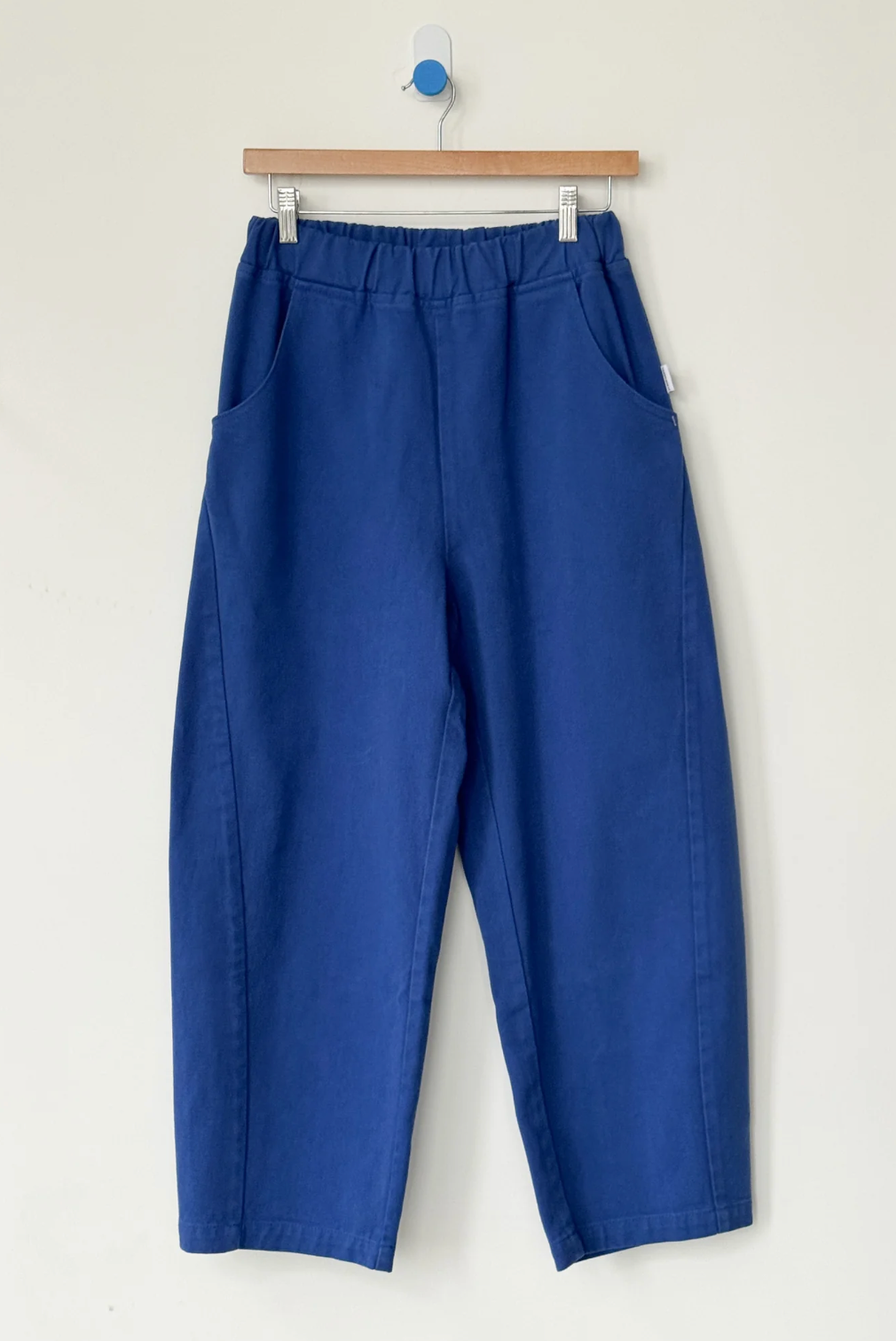 A pair of royal blue cropped pants with an elastic waistband and side pockets, hanging from a wooden hanger against a plain off-white wall.