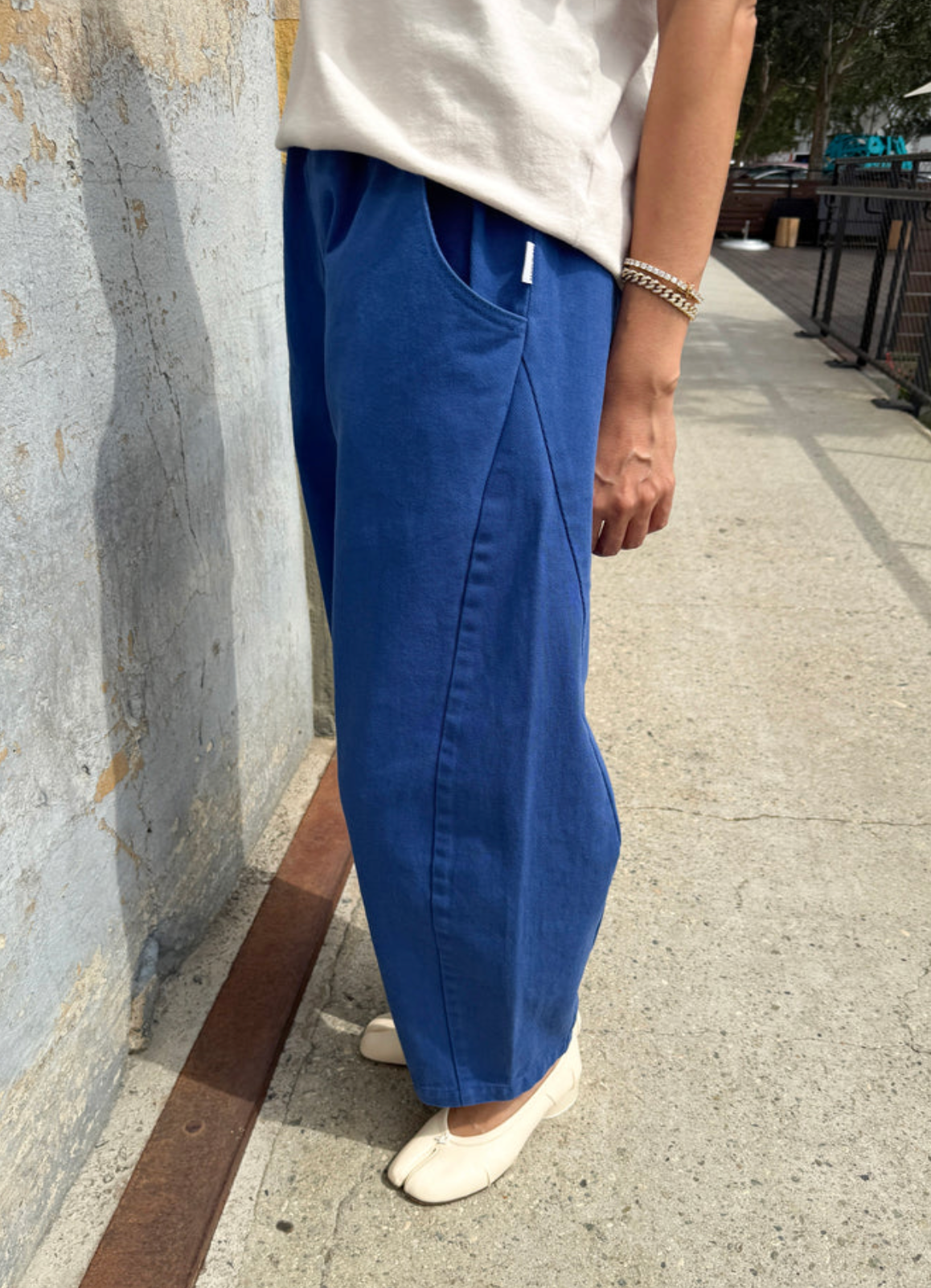 A person wearing wide-leg blue pants with a white t-shirt and white shoes, standing on a concrete outdoor walkway next to a textured gray wall.